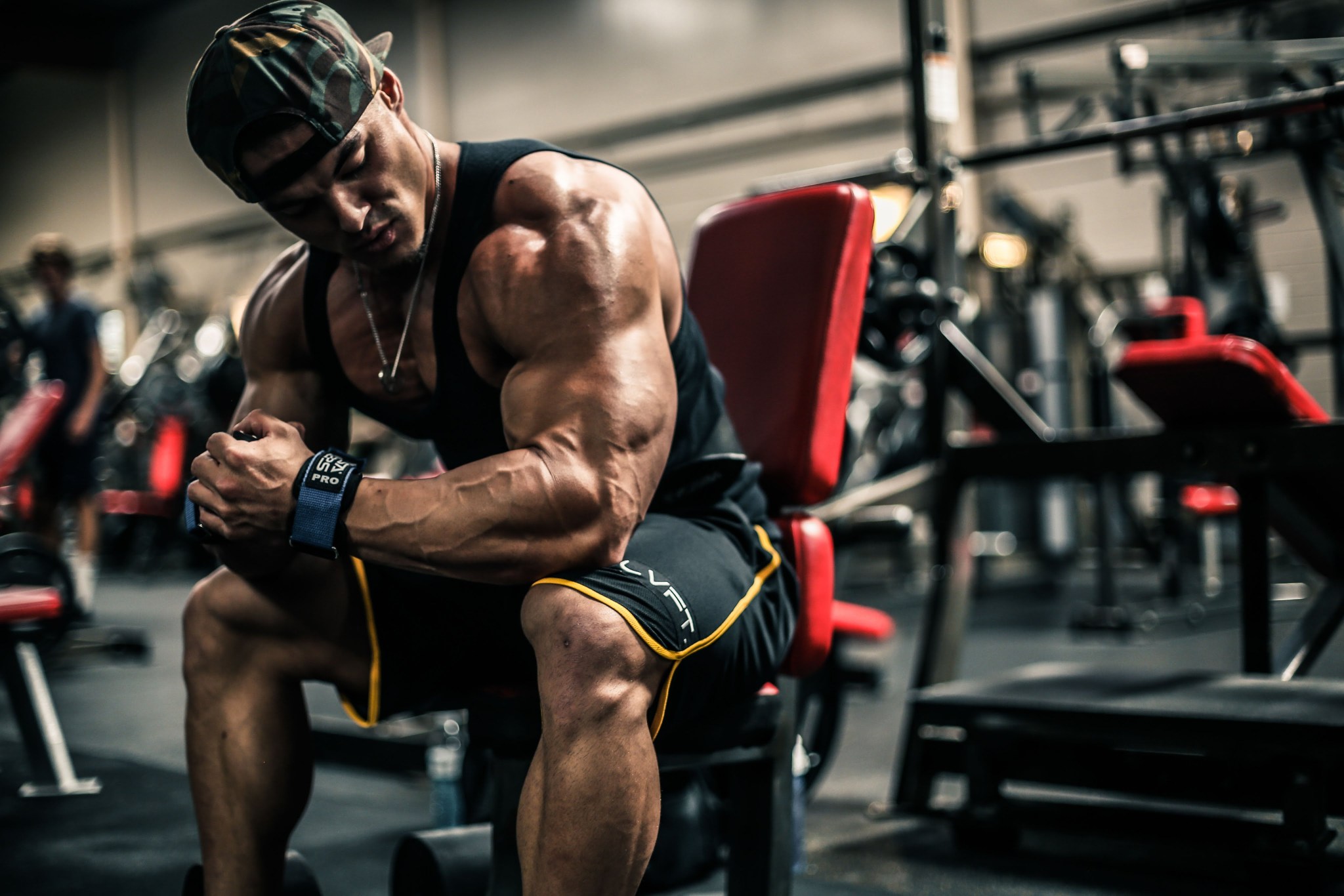 Muscular man in sportswear sits on a gym bench, flexes his arm and looks down, with weights in the background.