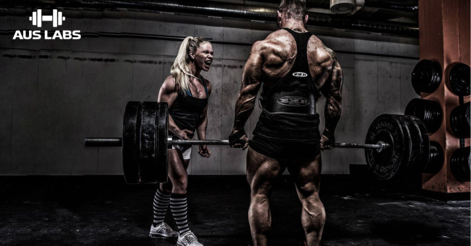A man lifts a heavy barbell in a gym as a woman encourages him; AUS LABS logo appears in the corner.