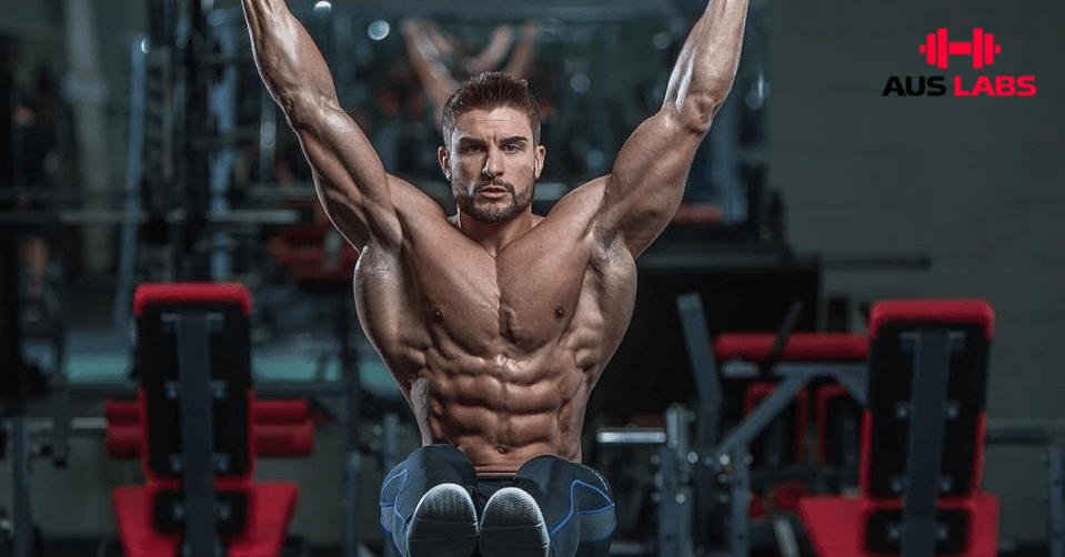 Muscular man doing a hanging leg raise in a gym with red equipment and AUS LABS logo in the background.
