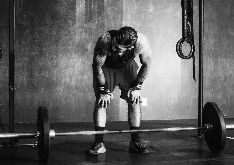 Man in gym gear rests over a barbell, hands on knees, with gymnastic rings visible behind him.