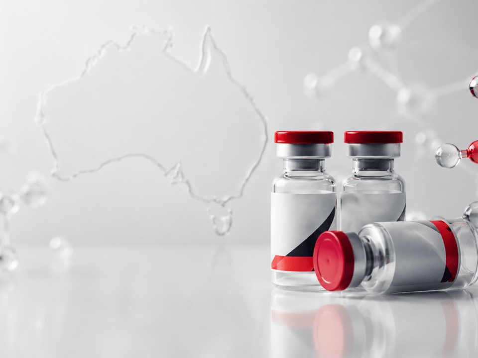 Three vaccine vials with red lids on a reflective surface, one lying down, beside a molecular model and Australia outline in back.